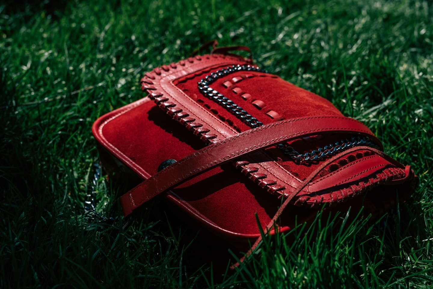 Borsa a tracolla in pelle scamosciata rossa appoggiata sull'erba verde lussureggiante.